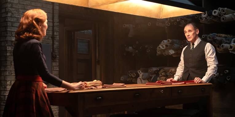 Zwei Menschen stehen in einem dunklen Raum in welchem Stoffe gelagert werden. Sie stehen sich jeweils am Kopfende eines Tisches gegenüber, über dem eine Lampe hängt, die die Szene ausleuchtet.